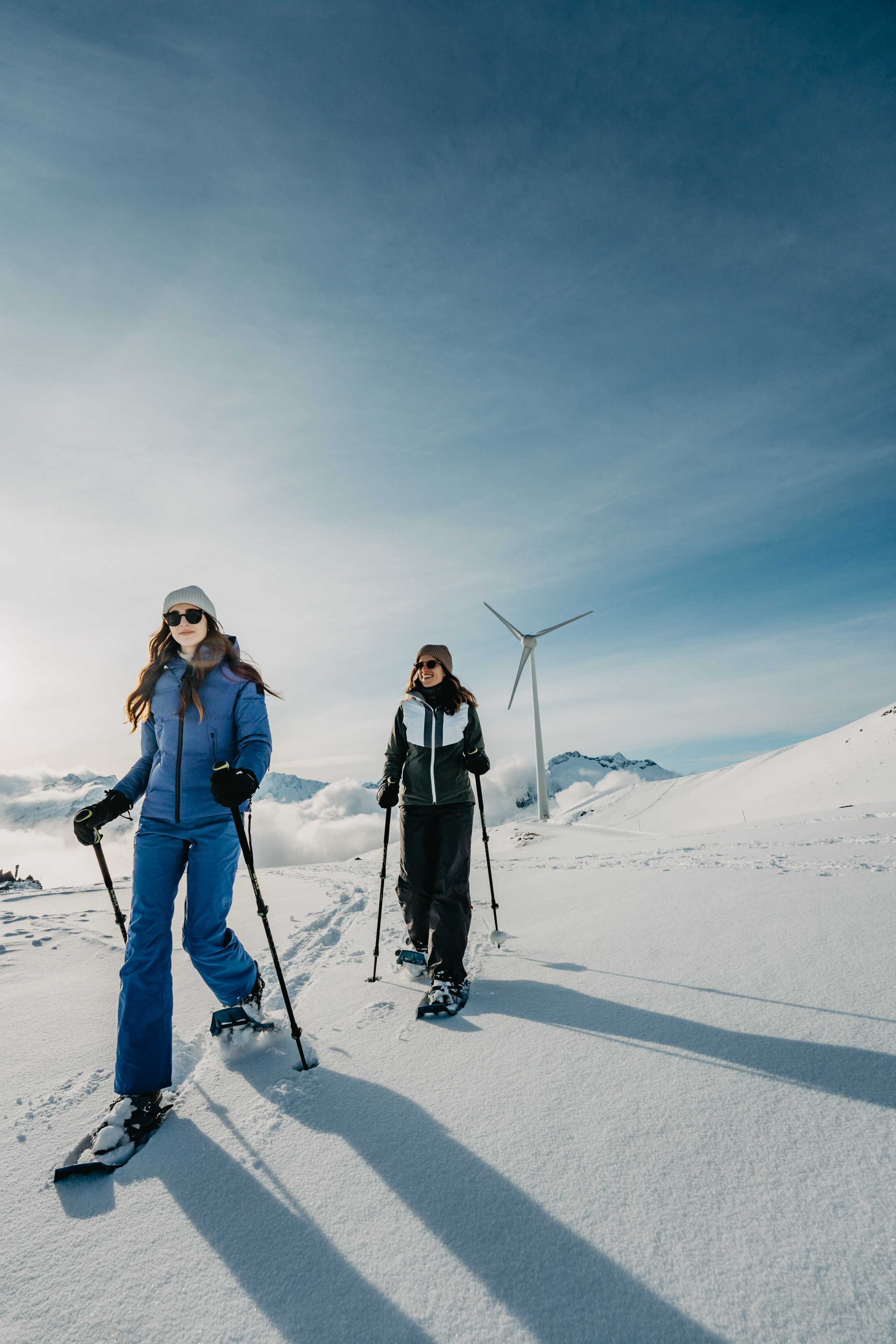 Two women snowshoeing in the Swiss alps. Wind turbines in the background.